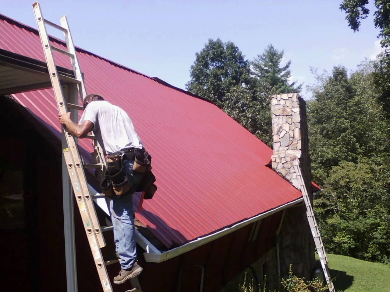 Roofer on a ladder beside a red metal roof and stone chimney — Soffit & Fascia Repair by Roofing Brothers in Groveton